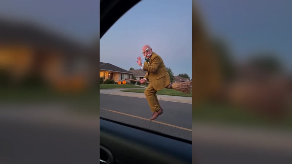 A man in a tweed suit rides a broomstick outside a car window on a suburban street at dusk